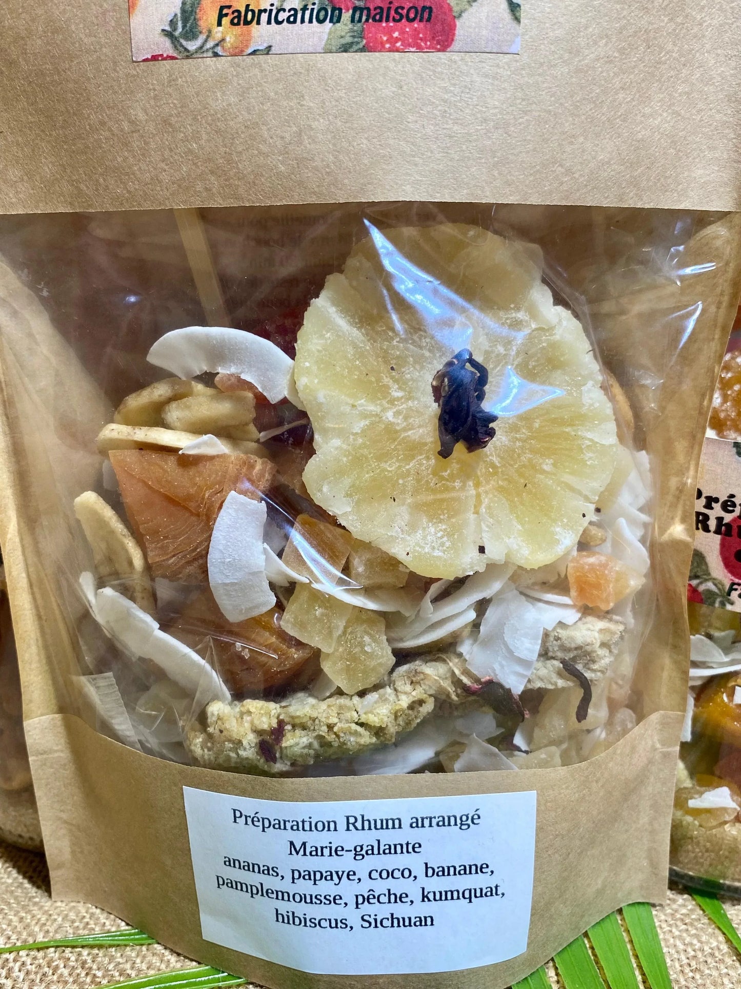 Gros plan des ingrédients du sachet de rhum arrangé La Marie‑Galante – ananas, papaye, noix de coco, banane, pomelo, pêche, kumquat, fleurs d’hibiscus séchées et poivre de Sichuan prêts à infuser dans du rhum blanc agricole
